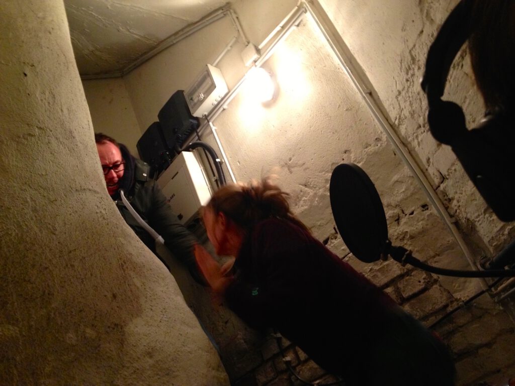 Zwei Personen in einem engen Kellerraum; daneben sind ein Mikrofon, ein Popfilter und ein Kopfhörer zu sehen.