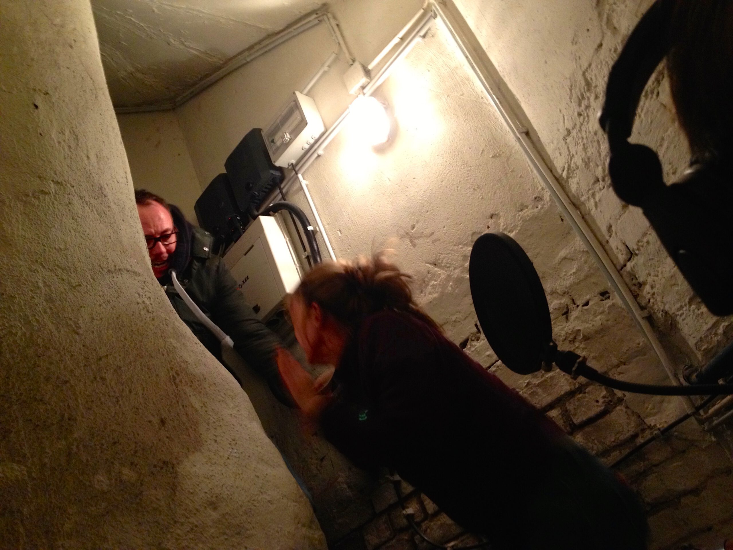 Zwei Personen in einem engen Kellerraum; daneben sind ein Mikrofon, ein Popfilter und ein Kopfhörer zu sehen.