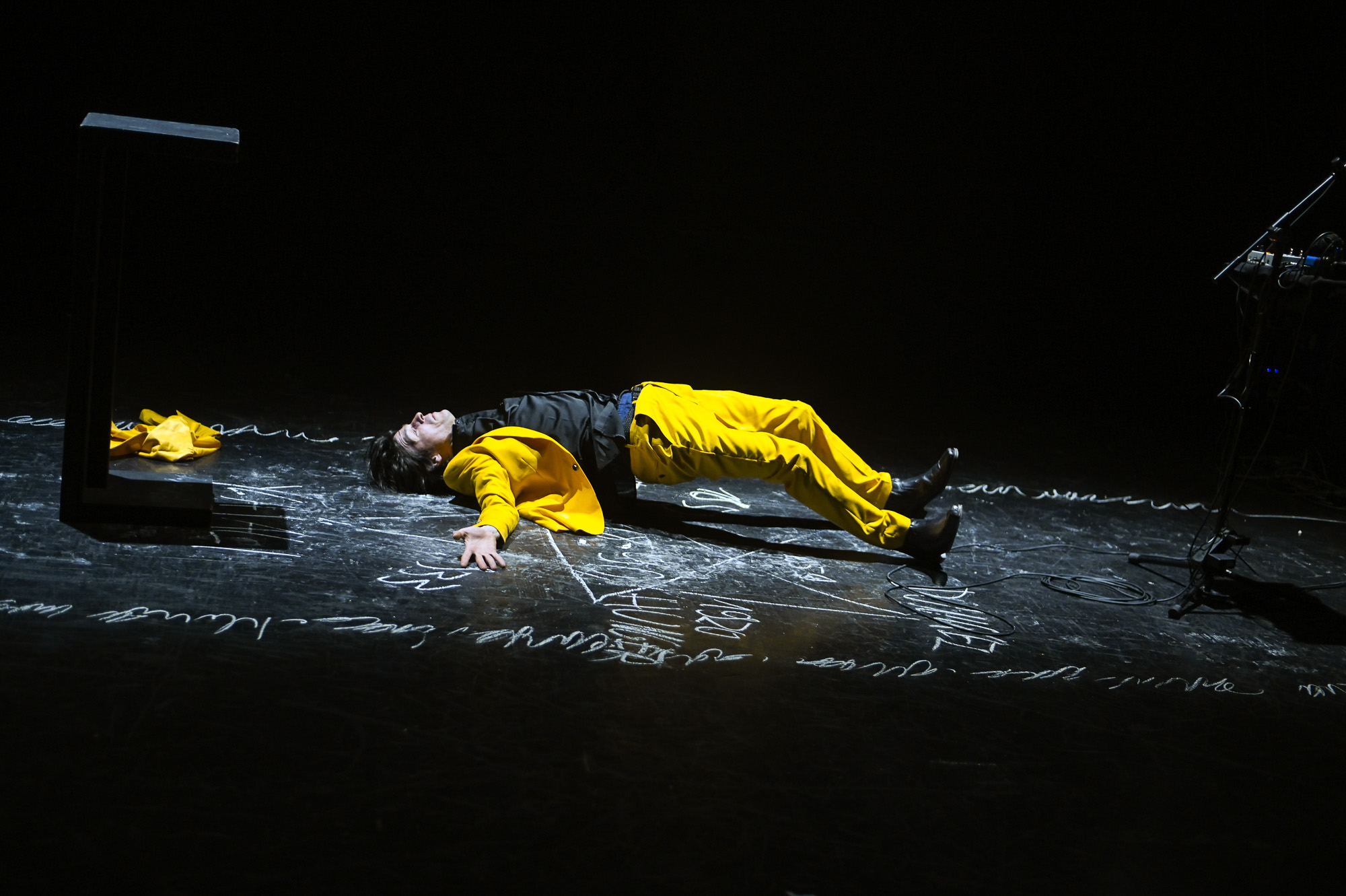Darsteller liegt in gelbem Anzug auf einer dunklen Bühne mit Kreideschrift auf dem Boden.