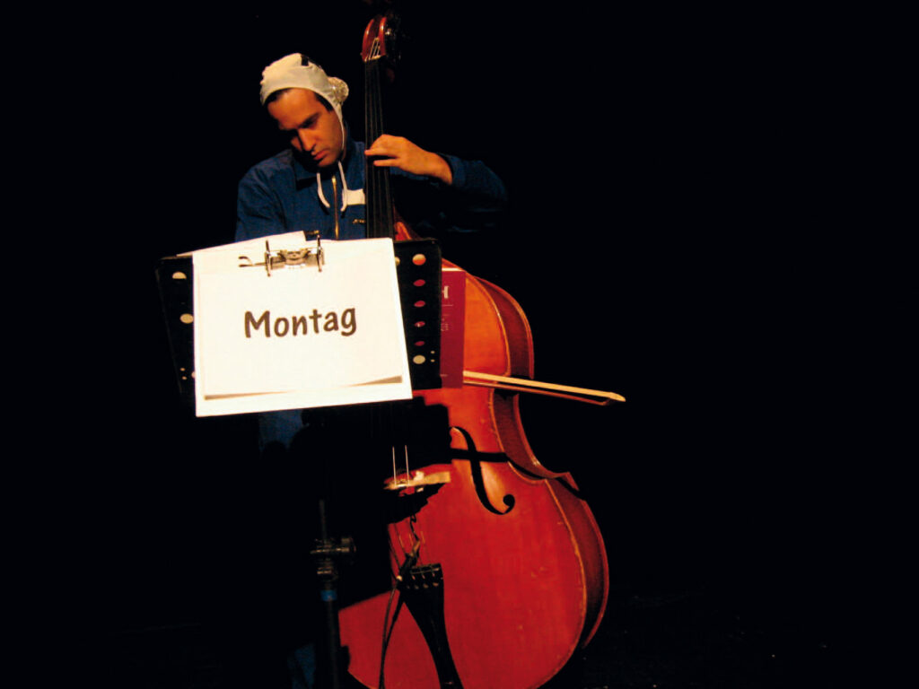 Ein Musiker steht mit einem Kontrabass auf einer dunklen Bühne; am Notenständer hängt ein Schild mit der Aufschrift „Montag“.